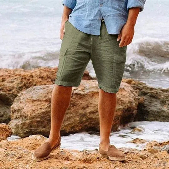 Uomo in camicia azzurra e bermuda verdi su spiaggia rocciosa, indossa mocassini marroni. Moda estiva casual, abbigliamento maschile mare.