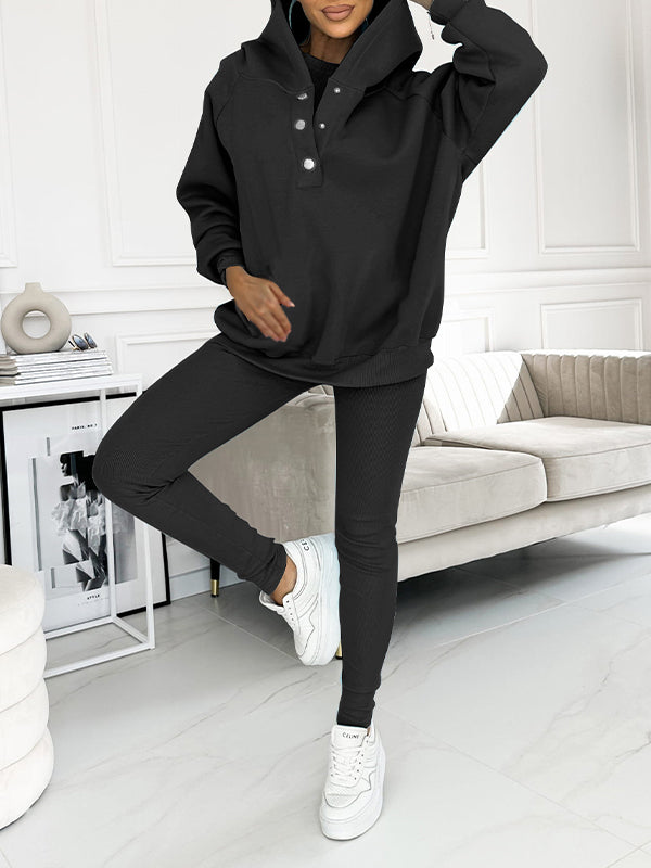 Donna in tuta sportiva nera, felpa con cappuccio e pantaloni aderenti, posa in salotto moderno. Scarpe da ginnastica bianche. Moda casual donna.
