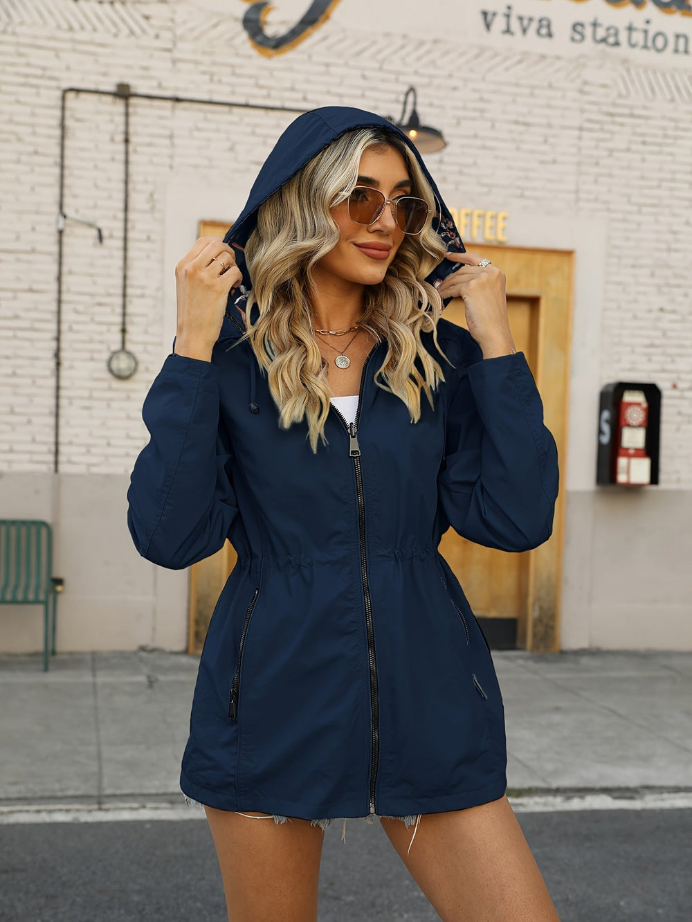 Donna con giacca a vento blu, cappuccio alzato, occhiali da sole, davanti a un edificio urbano. Moda autunnale, abbigliamento casual, stile urbano.