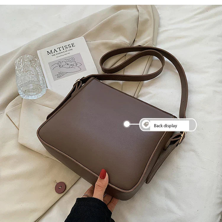 Borsa a tracolla marrone su tessuto beige, con libro d'arte e bicchiere. Design elegante, ideale per moda femminile e accessori di tendenza.