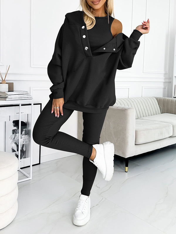 Donna in tuta nera elegante, con spalla scoperta e bottoni decorativi, posa in salotto moderno. Scarpe bianche completano il look casual-chic.