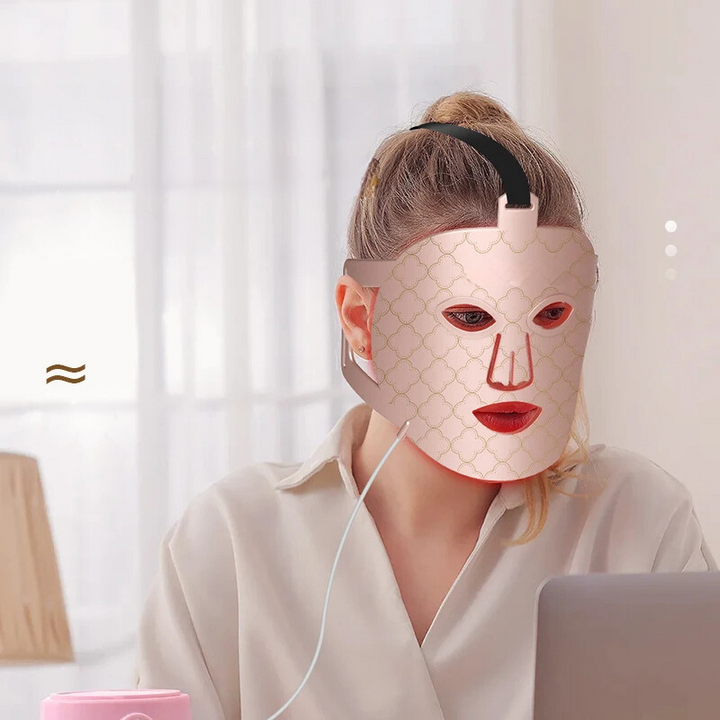 Donna con maschera LED per terapia della pelle, seduta al computer. Maschera viso bellezza, cura della pelle, trattamento anti-età, tecnologia LED.