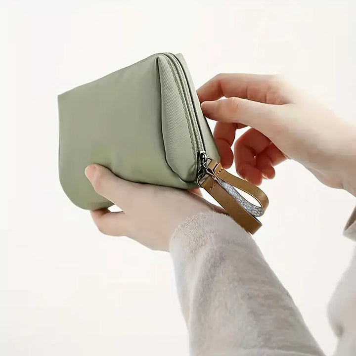 Mani che tengono un elegante astuccio verde in tessuto, chiuso con zip e cinturino in pelle. Accessorio moda, portafoglio compatto, design minimalista.