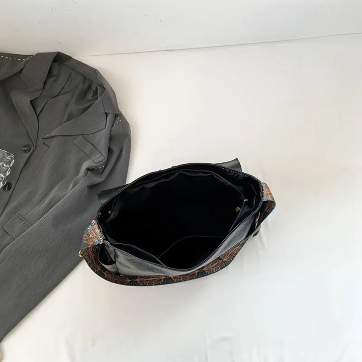 Borsa a tracolla nera in pelle con interno spazioso, accanto a giacca grigia su sfondo bianco. Accessorio moda elegante e versatile.