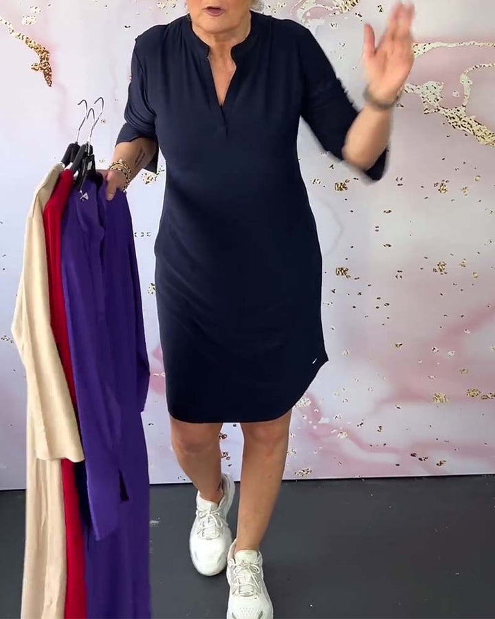 Donna in abito blu scuro, maniche a tre quarti, tiene abiti colorati su grucce. Sfondo rosa chiaro. Moda femminile, abbigliamento casual elegante.