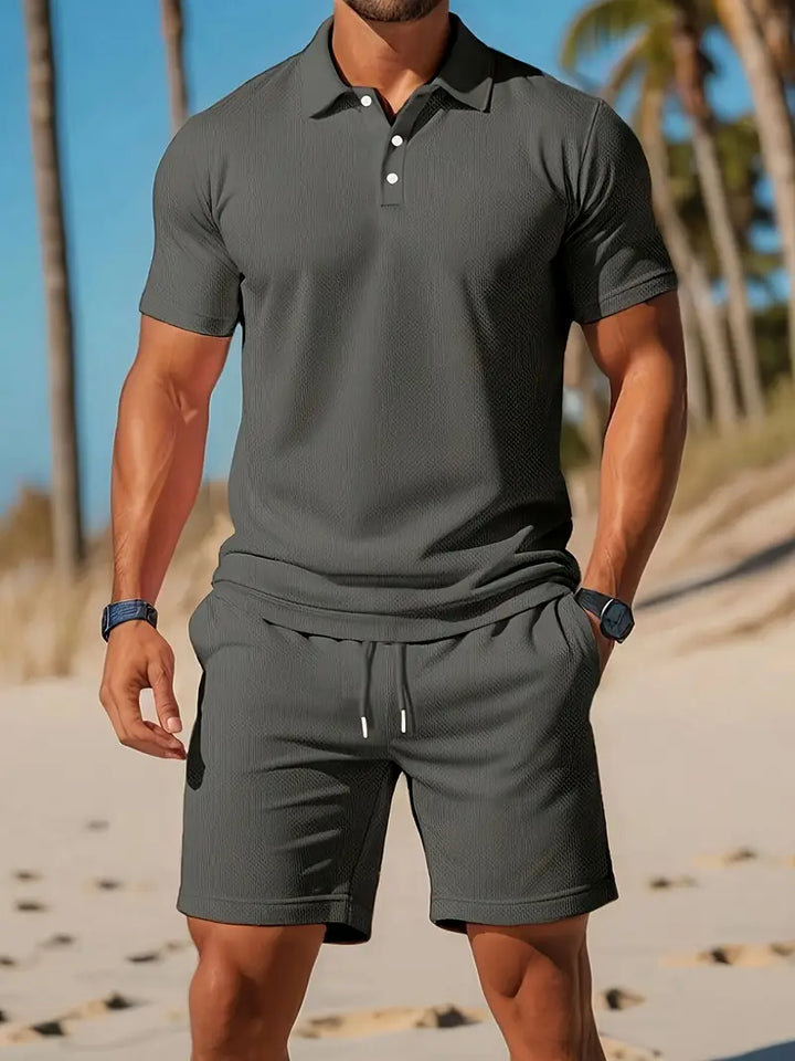 Uomo in spiaggia con polo e pantaloncini grigi, palme sullo sfondo. Abbigliamento estivo casual, moda maschile, look elegante e sportivo.