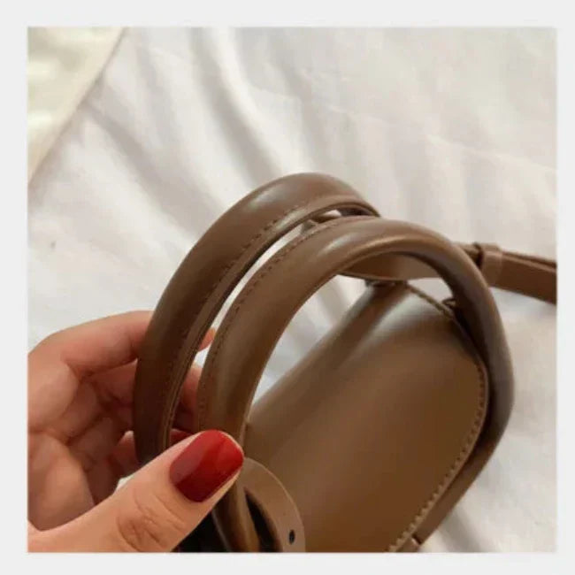 Mano femminile con smalto rosso tiene borsa a tracolla in pelle marrone su sfondo bianco. Accessorio moda elegante, stile minimalista.