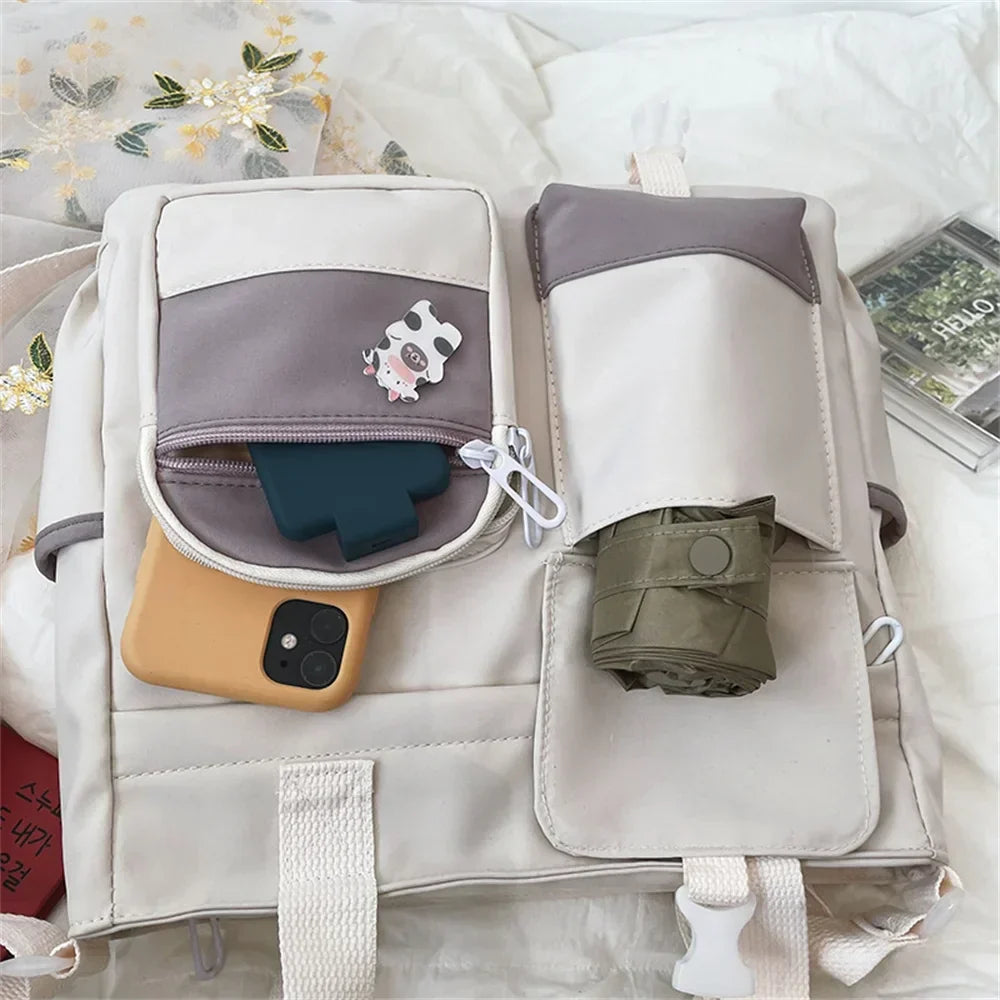 Zaino beige con tasche multiple, smartphone giallo, portachiavi mucca, ombrello pieghevole verde, su letto bianco. Accessori moda, organizzazione viaggio.