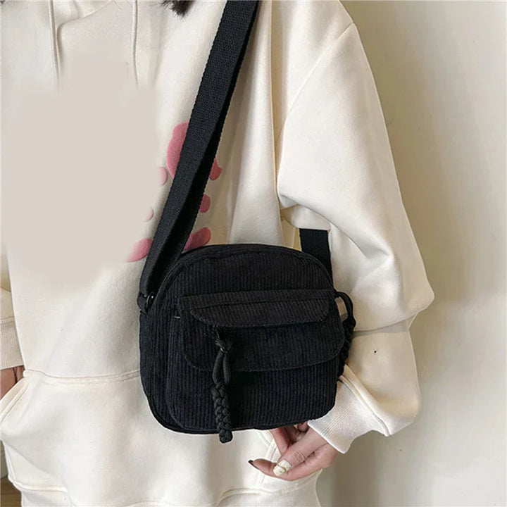Borsa a tracolla nera in velluto a coste, indossata su felpa bianca. Design compatto, ideale per moda casual e streetwear. Accessorio unisex.