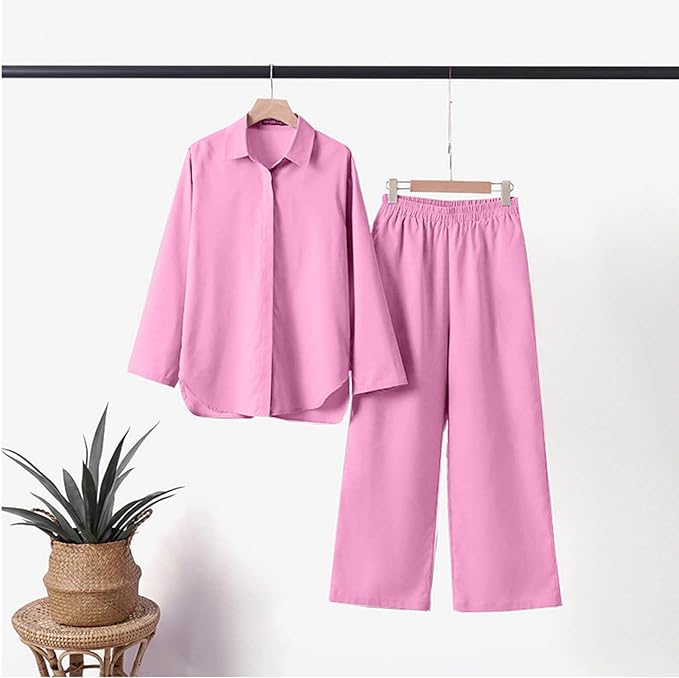 Completo pigiama donna rosa, camicia a maniche lunghe e pantaloni larghi, tessuto morbido, appeso su gruccia, pianta decorativa accanto.