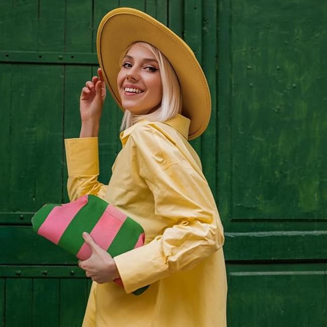 Donna sorridente con cappello giallo e camicia abbinata, tiene una borsa a righe rosa e verdi. Sfondo verde. Moda estiva, stile vivace.