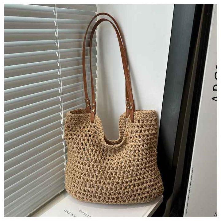 Borsa a tracolla in paglia intrecciata beige con manici in pelle marrone, appoggiata su una sedia vicino a una finestra con persiane grigie.