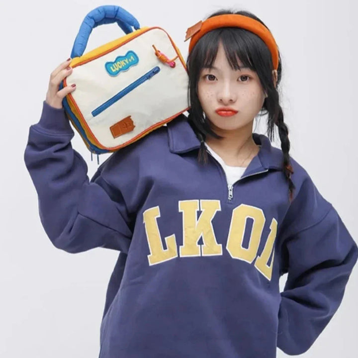 Ragazza con felpa blu "LKOL" e borsa colorata a mano, posa su sfondo bianco. Moda casual, accessori vivaci, stile giovanile.