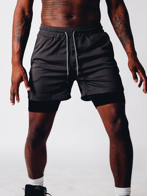 Uomo in pantaloncini sportivi neri, muscoloso, con tatuaggi sulle braccia, su sfondo bianco. Abbigliamento fitness, moda maschile, stile casual.