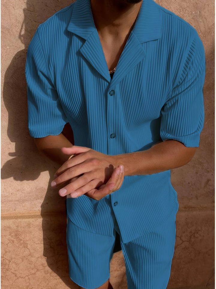 Uomo in abito estivo blu a righe, camicia a maniche corte e pantaloncini coordinati, in piedi contro un muro di pietra. Moda maschile estiva.