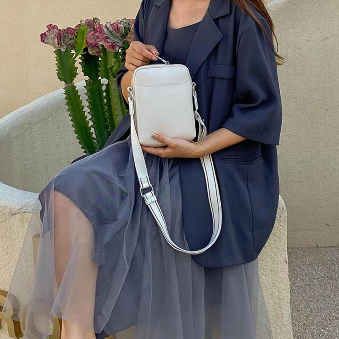 Donna seduta con abito blu e giacca, tiene una borsa a tracolla bianca. Sfondo con cactus. Moda elegante, accessori femminili, stile urbano.
