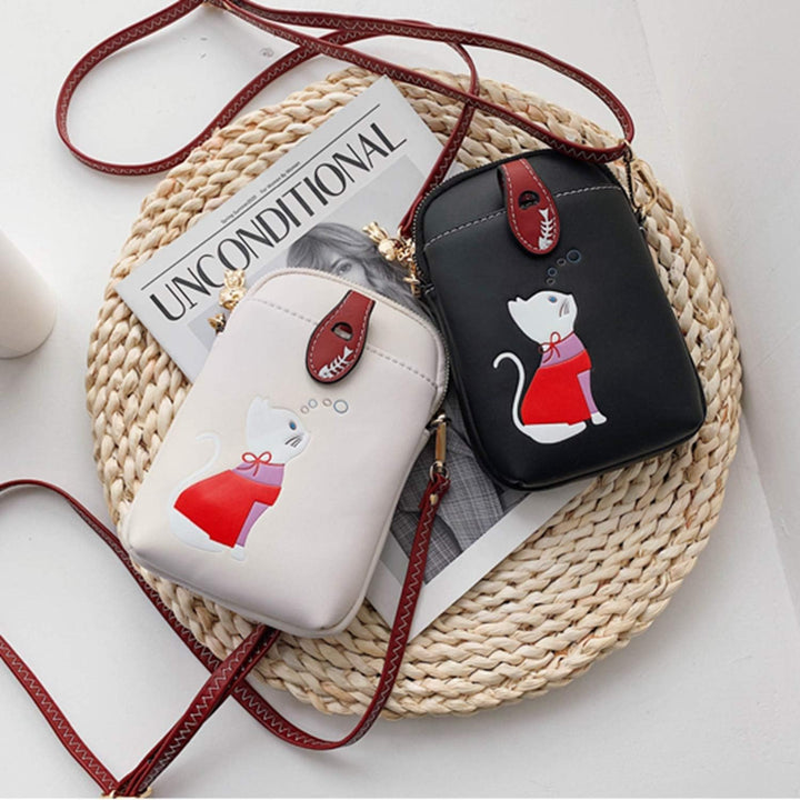 Due borse a tracolla in pelle, una bianca e una nera, con stampa di gatto, su un tappetino intrecciato accanto a una rivista. Accessori moda donna eleganti.