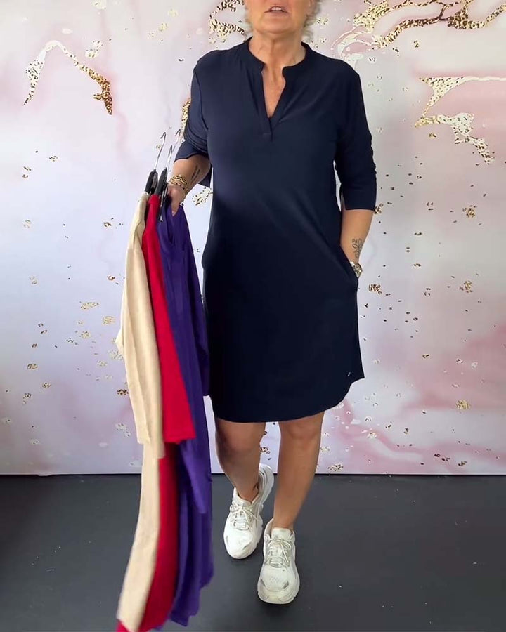 Donna con abito blu scuro, maniche a tre quarti, tiene abiti colorati su grucce. Sfondo decorativo rosa e bianco. Scarpe da ginnastica bianche.