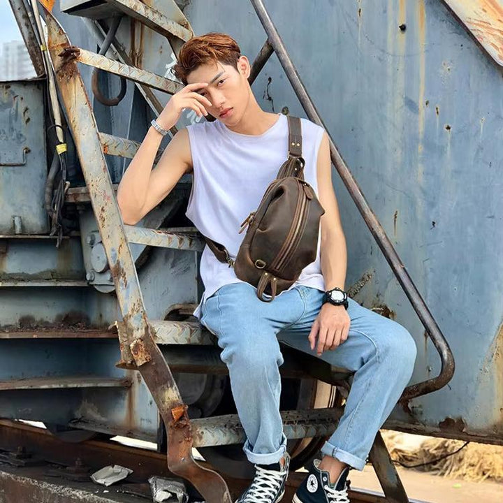 Giovane uomo seduto su scala metallica, indossa jeans, maglietta bianca senza maniche, borsa a tracolla marrone, orologio nero, scarpe Converse nere.
