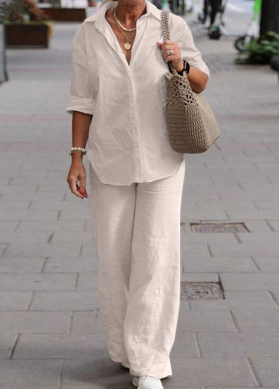 Donna con camicia bianca e pantaloni larghi, borsa intrecciata beige, cammina su marciapiede urbano. Moda casual elegante, abbigliamento estivo.