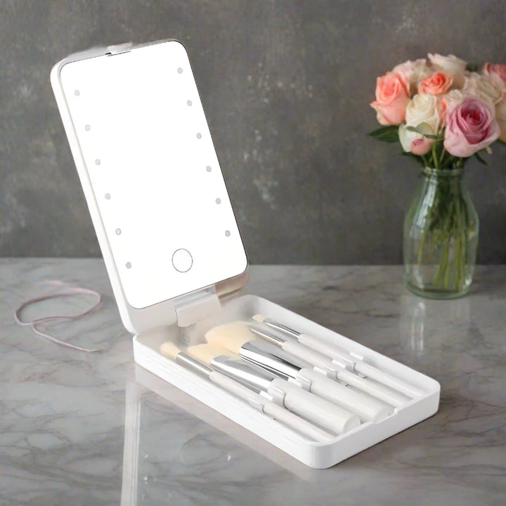 Set di pennelli da trucco con specchio LED portatile su tavolo di marmo, accanto a vaso di fiori. Accessori bellezza, trucco professionale.