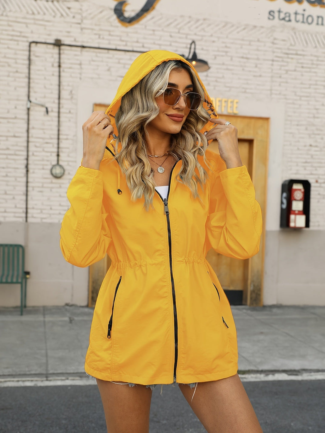 Giacca impermeabile gialla da donna con cappuccio, indossata da una modella all'aperto. Moda autunnale, stile casual, occhiali da sole.