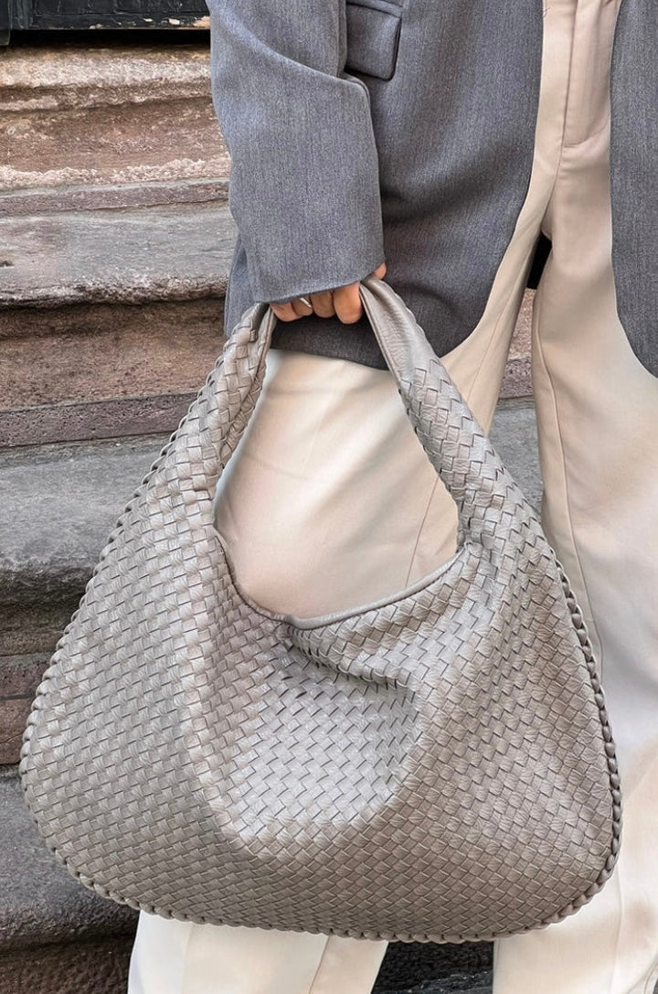 Borsa a spalla intrecciata in pelle grigia, indossata da una persona con giacca grigia e pantaloni bianchi. Accessorio moda elegante e moderno.