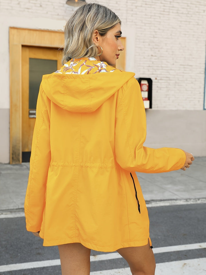 Donna con giacca impermeabile gialla, cappuccio e dettagli floreali, cammina in strada. Moda autunnale, abbigliamento casual, stile urbano.