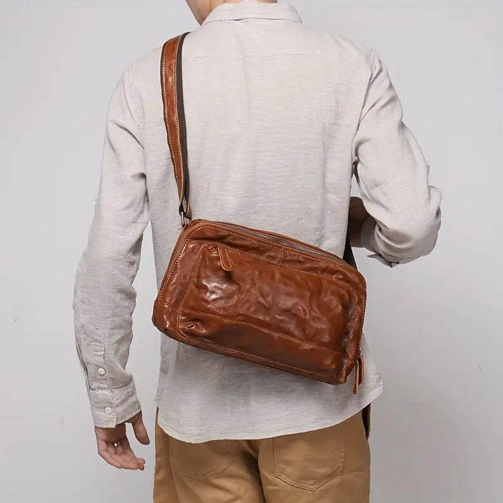 Borsa a tracolla in pelle marrone indossata da uomo con camicia beige e pantaloni marroni. Accessorio moda elegante e pratico per uso quotidiano.