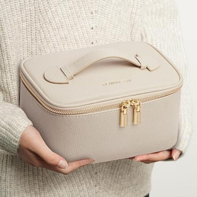 Mani che tengono un elegante beauty case beige in pelle con cerniera dorata e manico, ideale per cosmetici e accessori da viaggio.