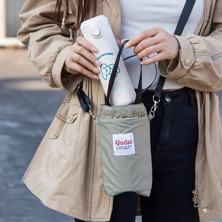 Donna con giacca beige tiene una borraccia bianca in una borsa a tracolla grigia. Accessorio pratico per bevande, ideale per viaggi e attività all'aperto.