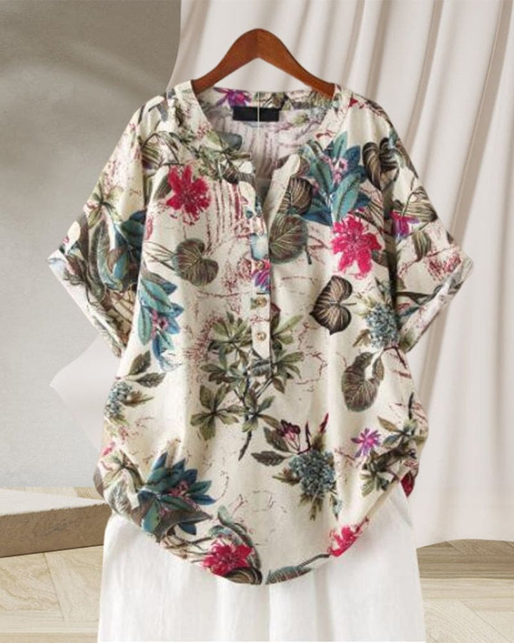 Camicia donna floreale in cotone, maniche corte, colletto a V, bottoni, sfondo bianco, design casual estivo, appesa su gruccia di legno.