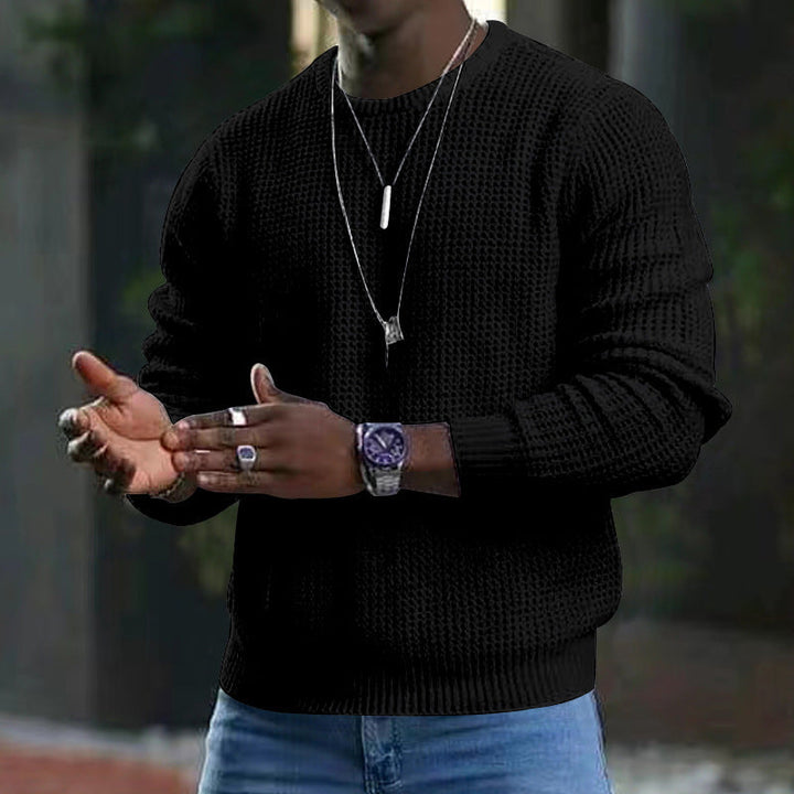 Maglione nero a maglia grossa indossato da uomo, abbinato a jeans blu. Accessori: orologio sportivo, collane e anelli argento. Moda casual elegante.