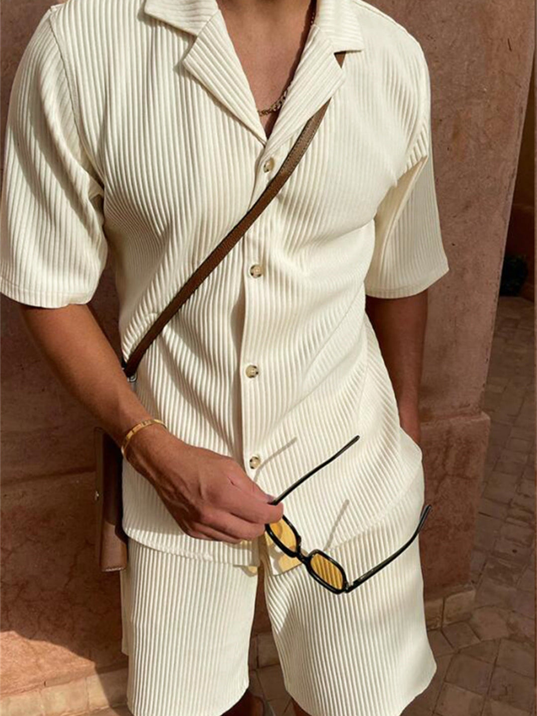Uomo con completo estivo beige a righe, camicia a maniche corte e pantaloncini coordinati, occhiali da sole in mano, stile casual elegante.