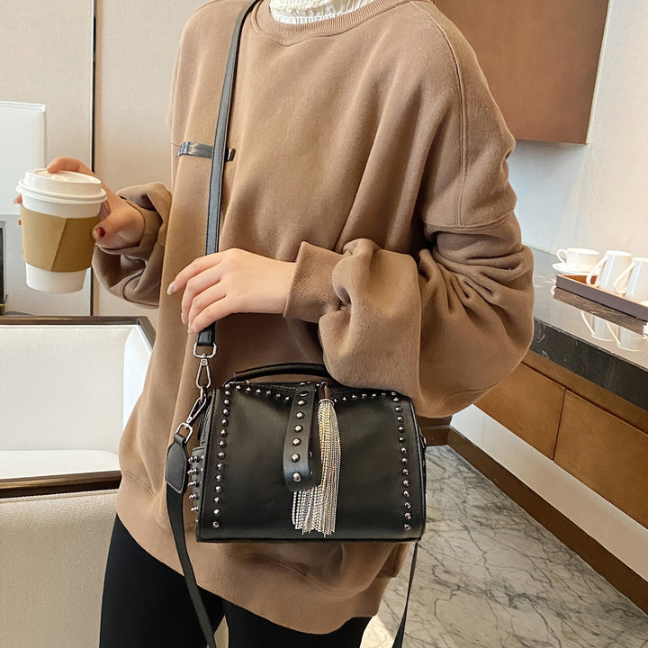 Donna con borsa a tracolla nera con borchie e frange, indossa maglione marrone, tiene bicchiere di caffè. Accessorio moda elegante e casual.