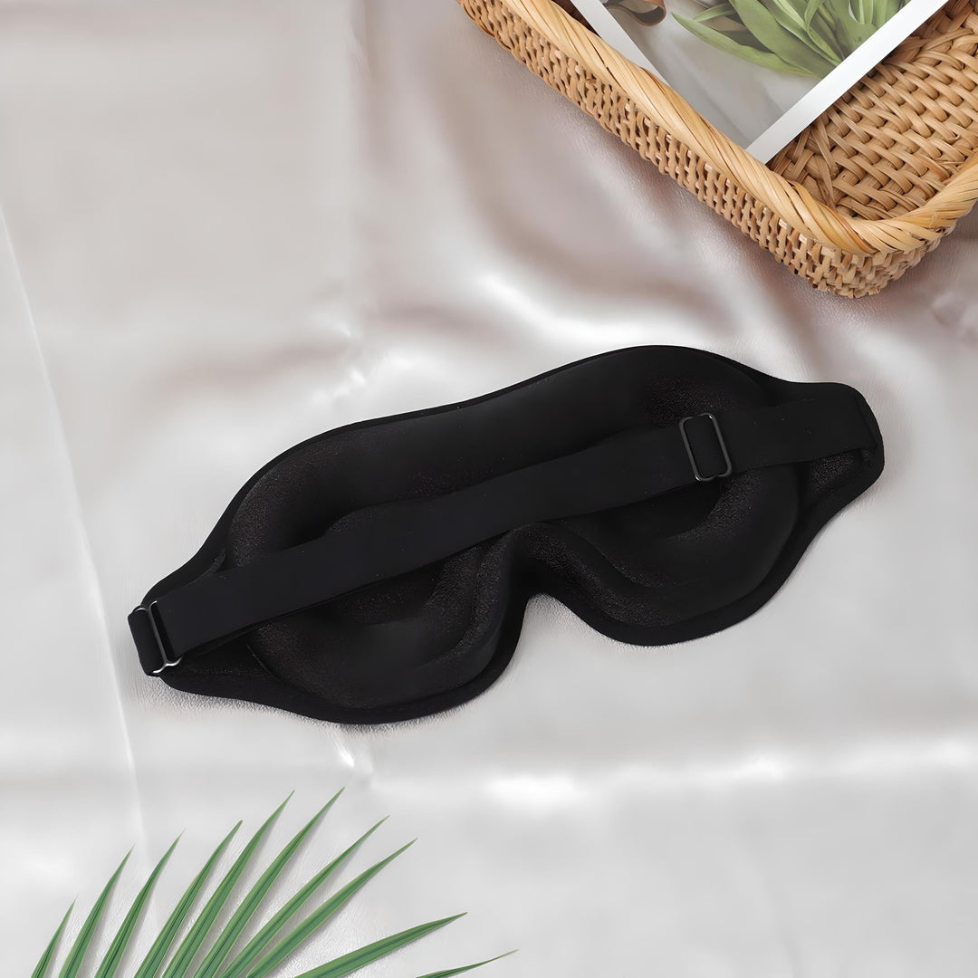 Maschera per dormire nera su tessuto bianco, con cinturino regolabile. Accessorio per il sonno, relax, viaggio. Sfondo con foglia e cesto.