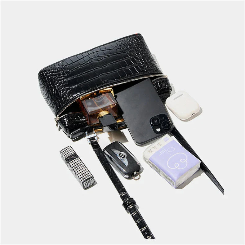 Borsa a tracolla nera in pelle con texture coccodrillo, contenente smartphone, profumo, chiavi, rossetto, auricolari e fazzoletti.