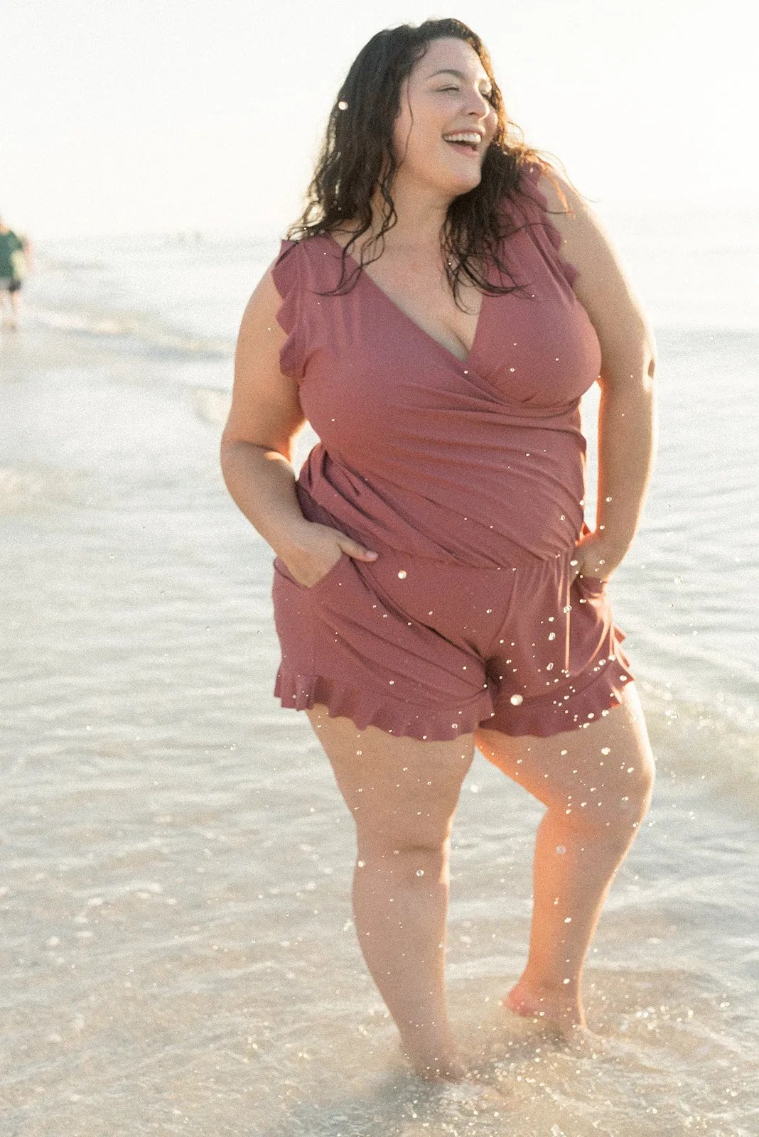 Donna sorridente in spiaggia con tuta rosa, moda mare taglie forti, estate, abbigliamento comodo, acqua, sole, felicità, stile casual.