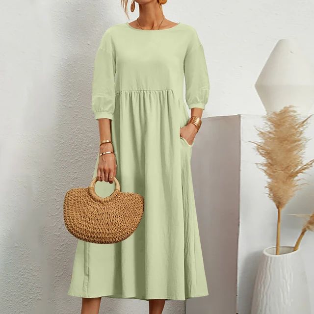 Abito lungo verde chiaro con maniche a tre quarti, tasche laterali, indossato da donna. Accessori: borsa in paglia, bracciali dorati.