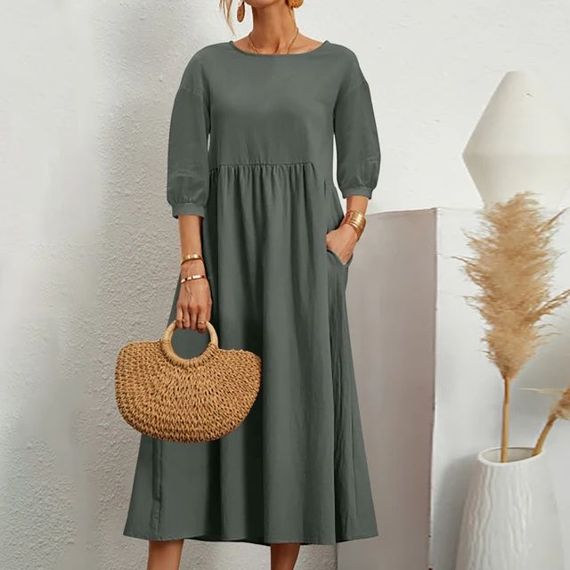 Abito lungo verde oliva con maniche a tre quarti, tasche laterali, indossato da donna. Accessori: borsa in paglia, bracciali dorati.
