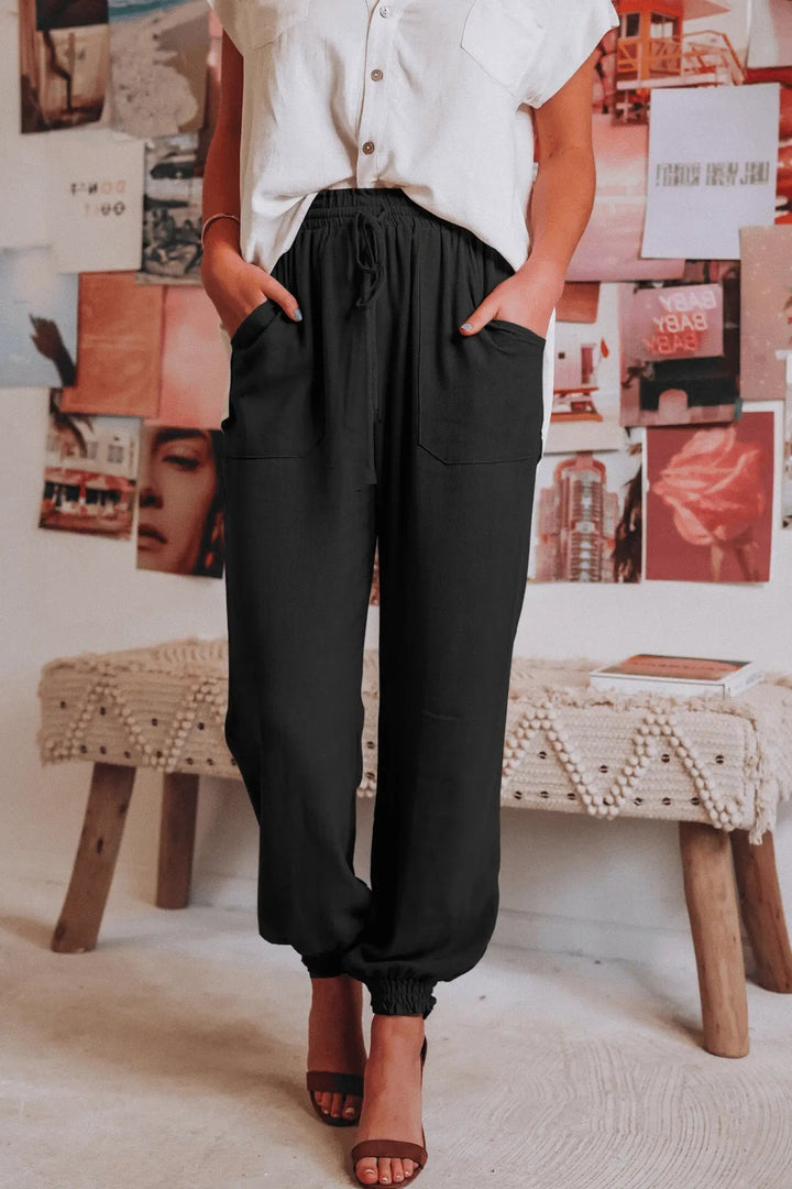 Donna in pantaloni neri larghi e camicia bianca, in piedi su un tappeto beige, sfondo con poster artistici. Moda casual, abbigliamento donna elegante.