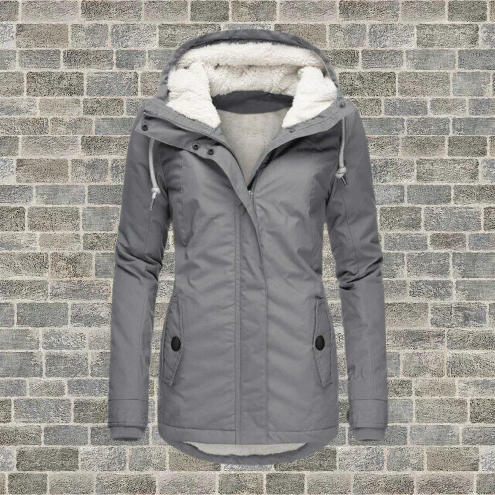 Giacca invernale grigia da donna con cappuccio foderato in pelliccia sintetica, chiusura a zip, tasche frontali, su sfondo di mattoni grigi.