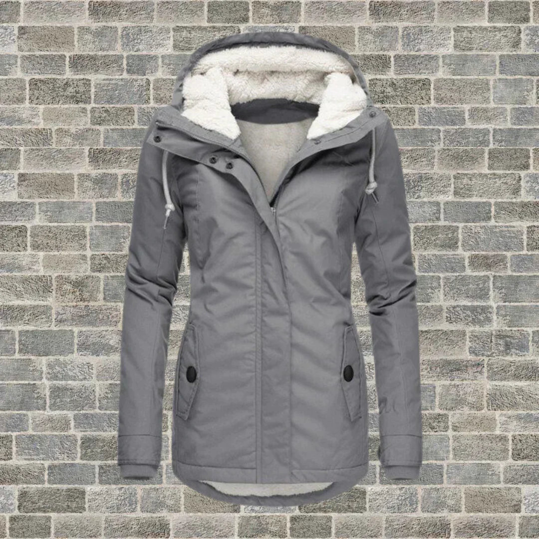 Giacca invernale grigia da donna con cappuccio foderato in pelliccia sintetica, chiusura a zip, tasche frontali, su sfondo di mattoni grigi.