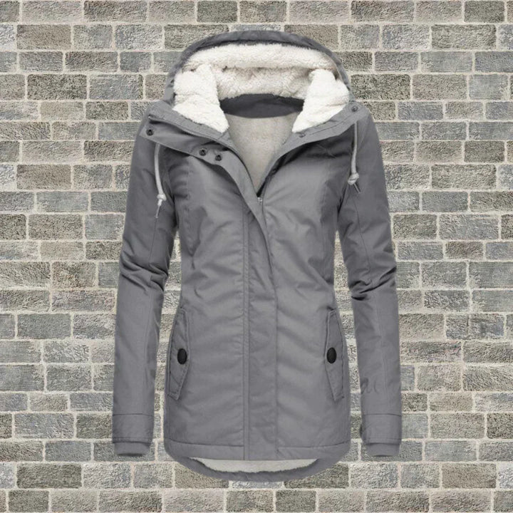 Giacca invernale grigia da donna con cappuccio foderato in pelliccia sintetica, chiusura a zip, tasche frontali, su sfondo di mattoni grigi.