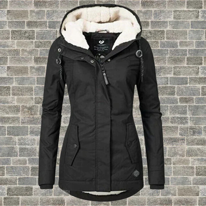 Giacca invernale nera da donna con cappuccio foderato in pelliccia sintetica, chiusura a zip, tasche frontali, su sfondo di mattoni grigi.