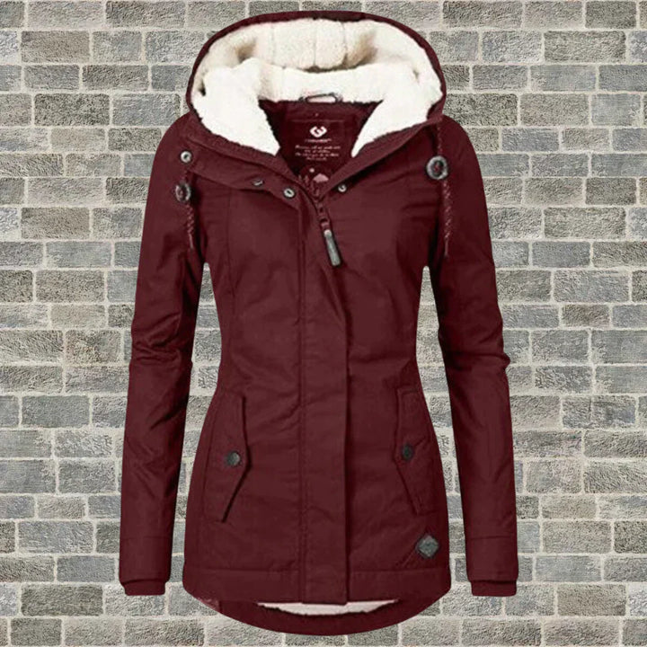 Giacca invernale da donna bordeaux con cappuccio foderato in pelliccia sintetica, chiusura a zip, bottoni decorativi, su sfondo di mattoni grigi.