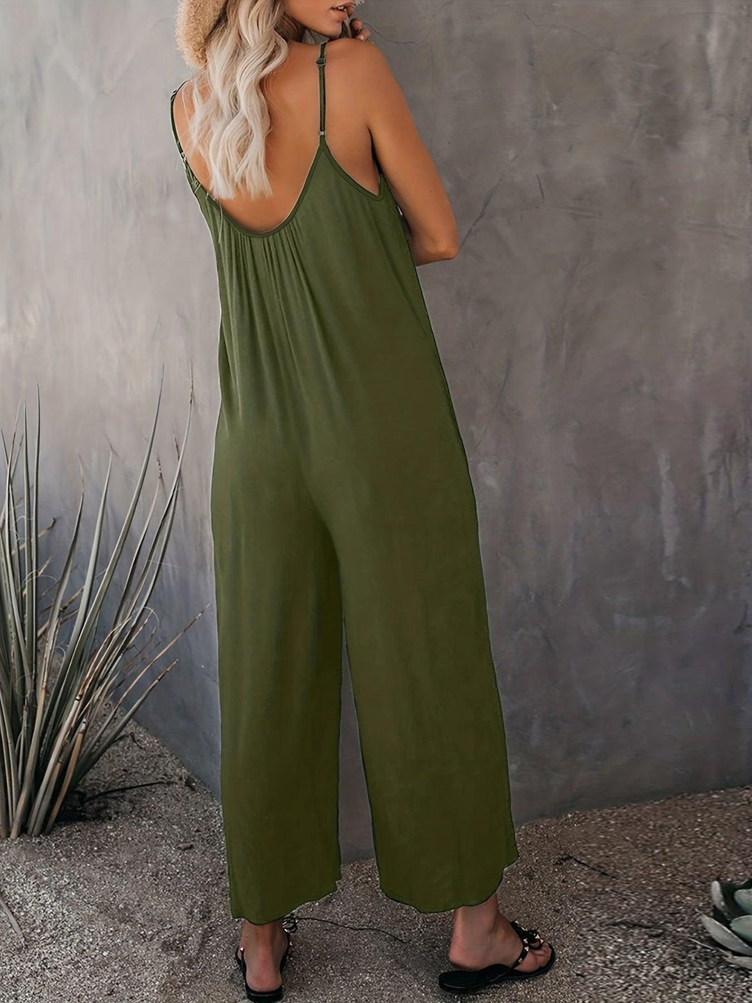 Donna con tuta verde oliva senza maniche, sandali neri, cappello di paglia, sfondo grigio. Moda estiva casual, abbigliamento femminile trendy.