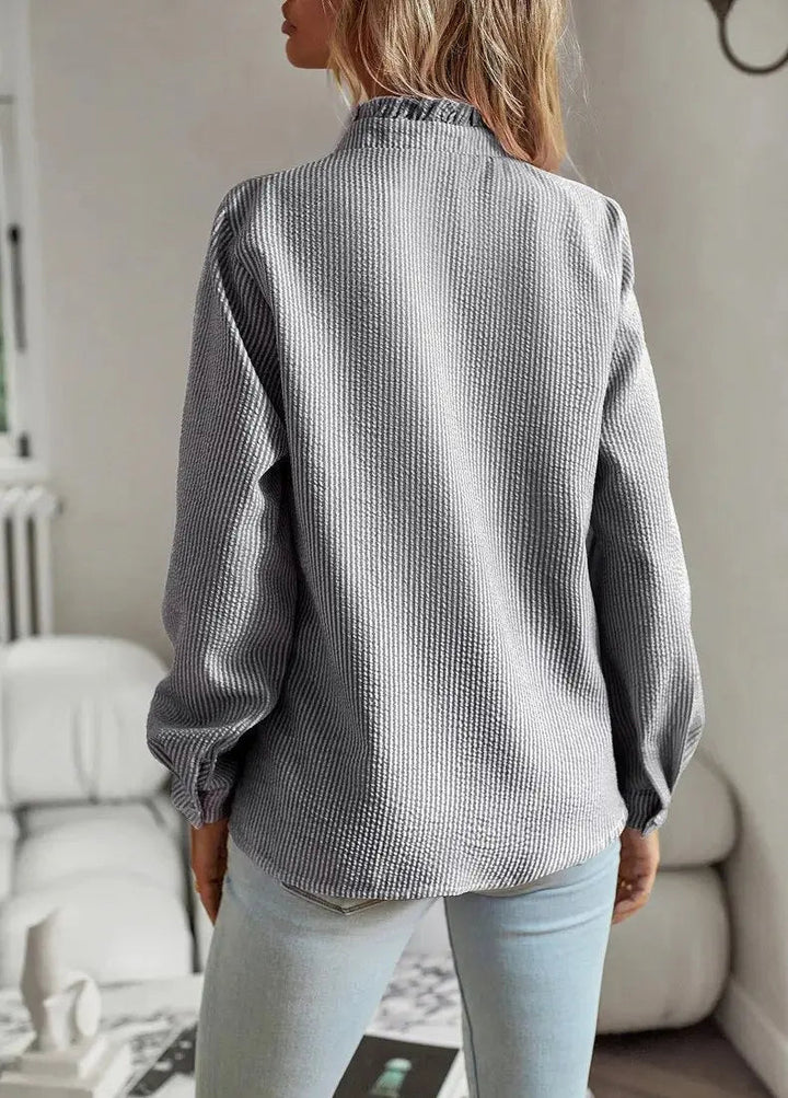 Donna con camicia a righe grigie, maniche lunghe, collo alto, in un ambiente interno moderno. Moda casual, abbigliamento femminile autunnale.