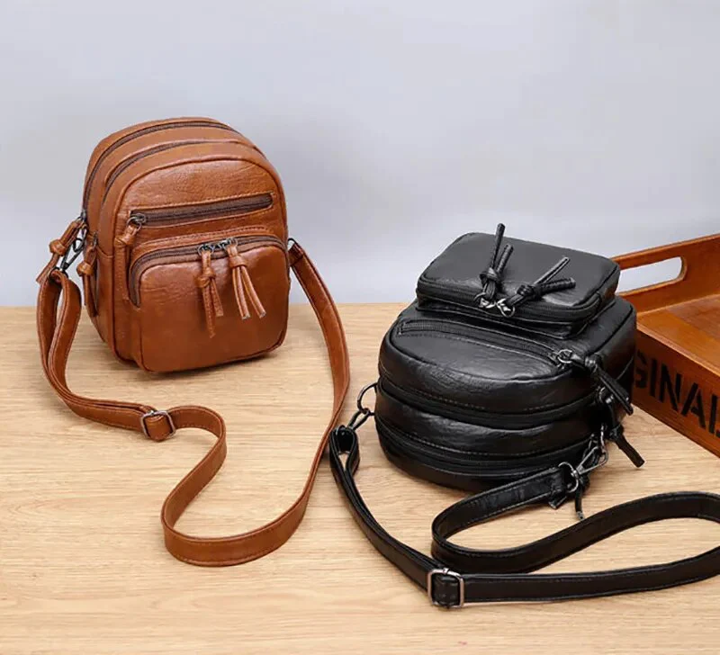 Due zaini in pelle, uno marrone e uno nero, con cinghie regolabili, su un tavolo di legno. Design compatto, ideale per viaggi e uso quotidiano.