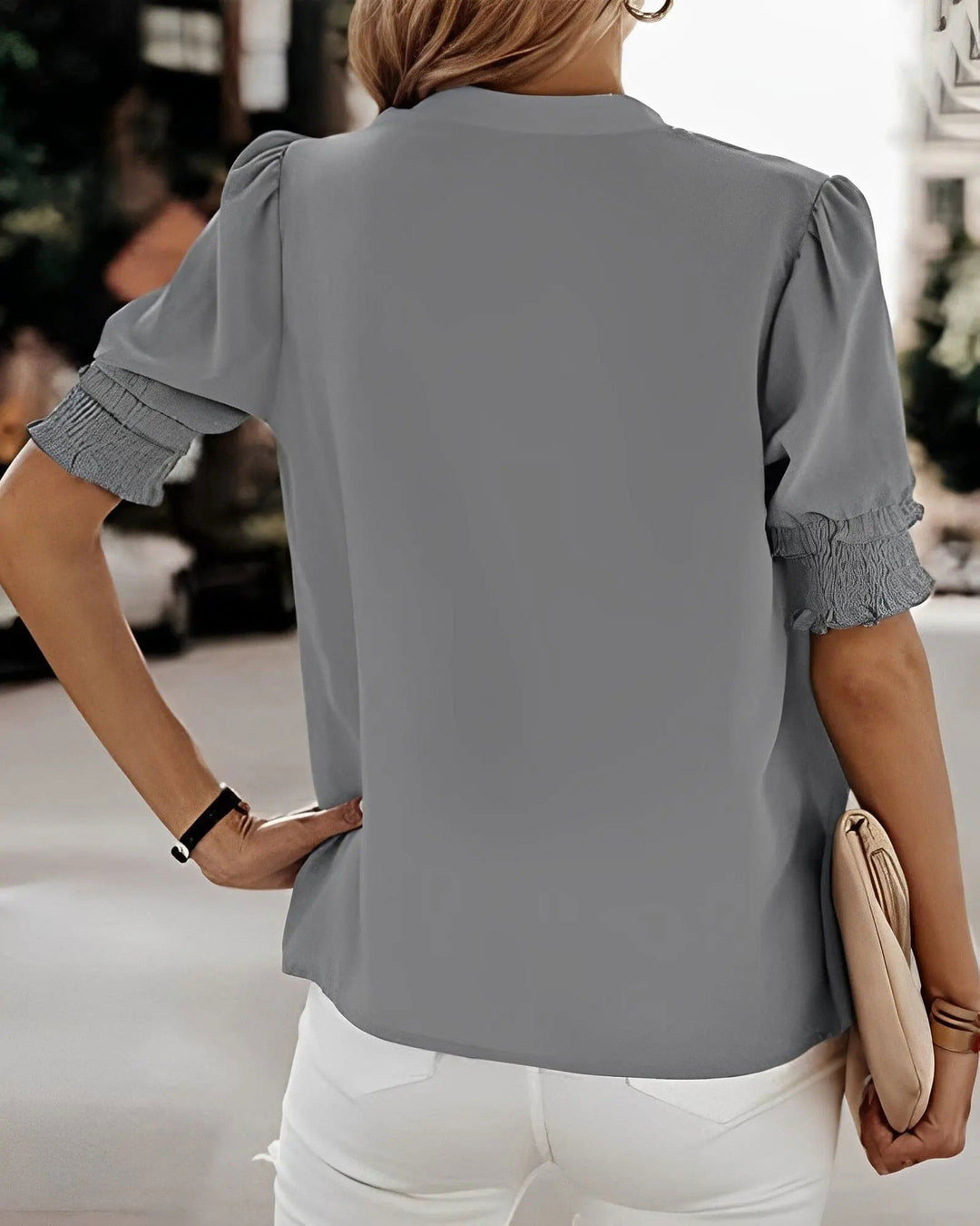 Donna con camicetta grigia a maniche corte e pantaloni bianchi, borsa beige, sfondo urbano. Moda estiva casual, abbigliamento femminile elegante.